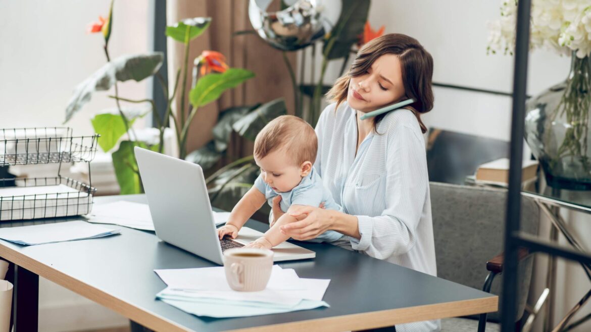Young working parent with child on lap while working on laptop and phone to ear